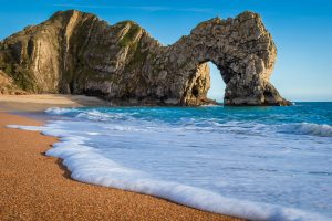 Durdle Door Beach - Waves lap the terracotta sand/shingle coast of one of the most famous of Dorset Beaches with its arch-shaped rock formation in the distance