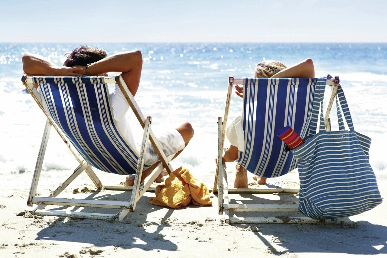 A couple relaxing on the beach on deck chairs from behind