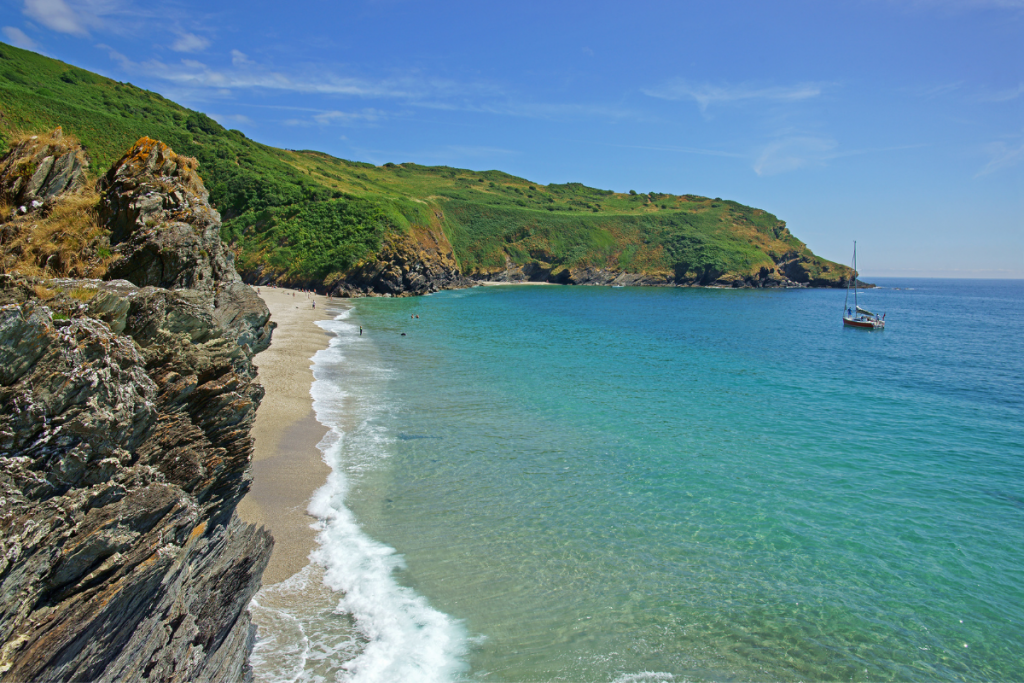 South Cornwall beach on a sunny day. Turquoise sea lapping golden sand against a grassy cliff backdrop