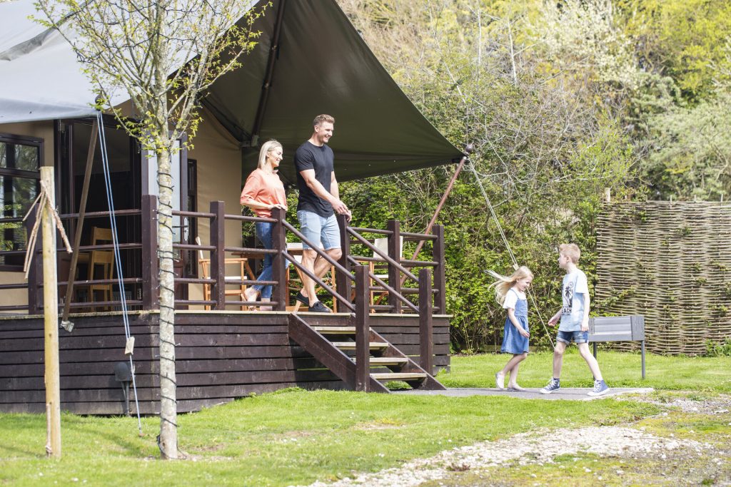 A young couple standing on the veranda of a wooden safari lodge whle their two children play together on the grassy outdoor area